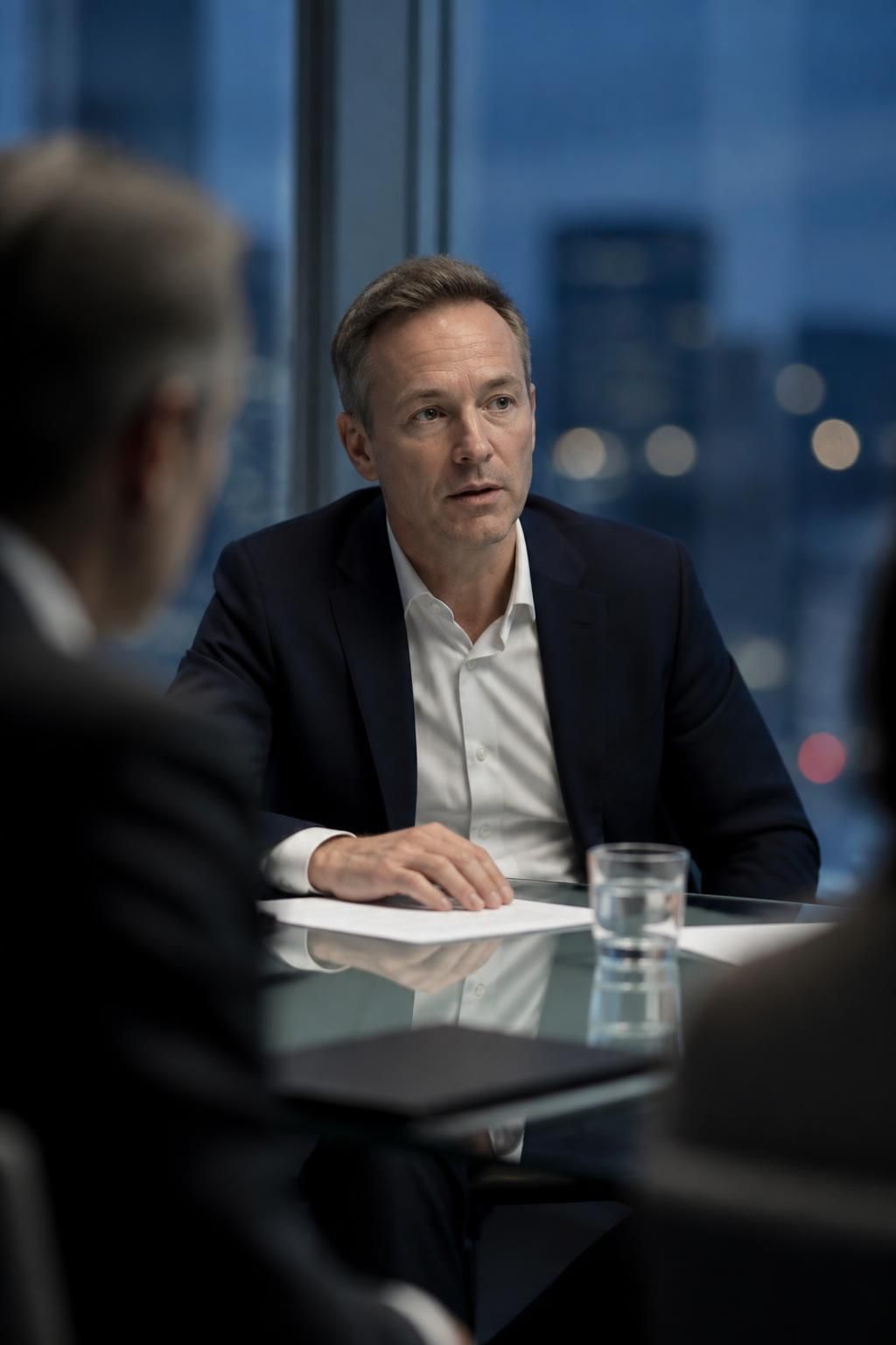 A professional in a navy suit at a glass meeting table, pausing mid-sentence